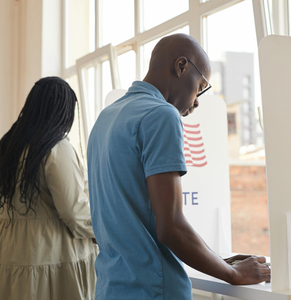 Photo of two Black voters in voting booth. Woman on left in green dress. Man on right in blue shirt. On the voting booth partition there is an American flag icon and the word "VOTE"